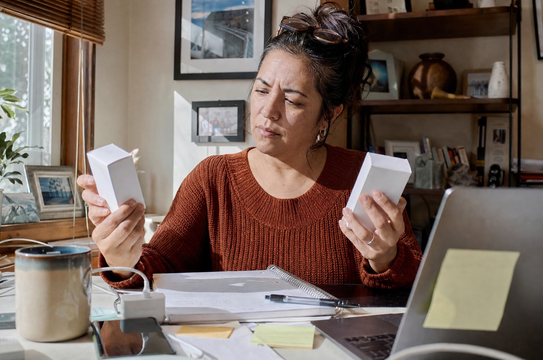 Woman in her late forties at a cluttered home-office desk comparing the ingredient lists on two skincare boxes next to an open notebook