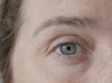 Extreme close-up of one eye and the full eyebrow of a 49-year-old woman with freckles and naturally sparse lashes, no mascara, in soft window daylight