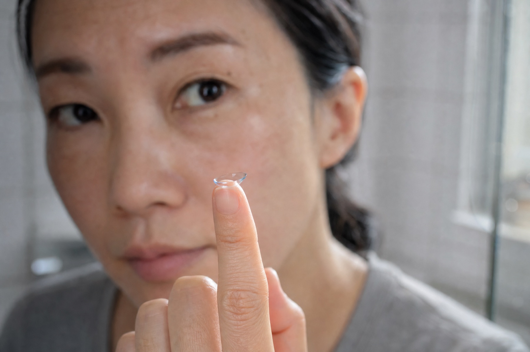 Soft contact lens balanced on a fingertip in front of a bathroom mirror, a woman's eye and eyebrow out of focus behind it