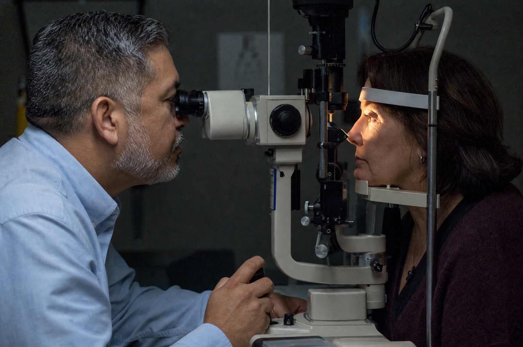 Male optometrist in his early fifties examining a woman's eye with a slit lamp in a dim exam room, her eyebrow and lashes visible in the lamp's glow