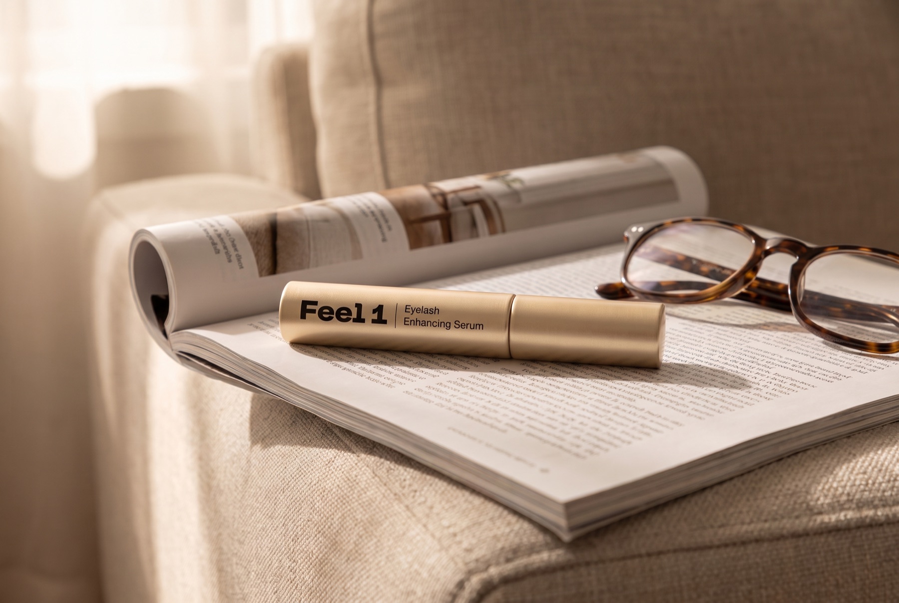 Gold Feel1 eyelash serum tube lying on an open magazine on a sofa armrest next to a pair of reading glasses