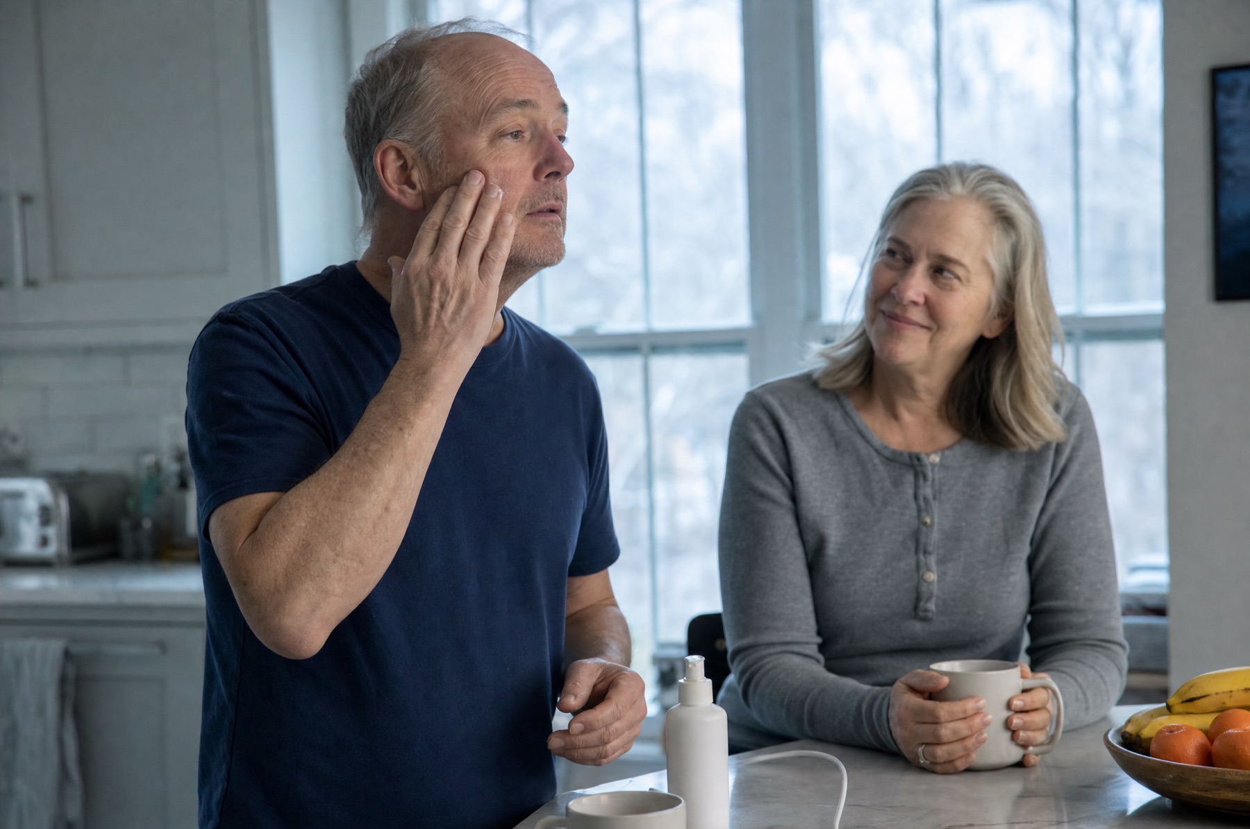 Man of about sixty rubbing moisturizer into his cheek at the kitchen counter in early morning light while his wife watches