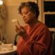 Woman in her fifties reading the back of a small skincare tube at her bathroom sink in the evening, a jar of cream beside her