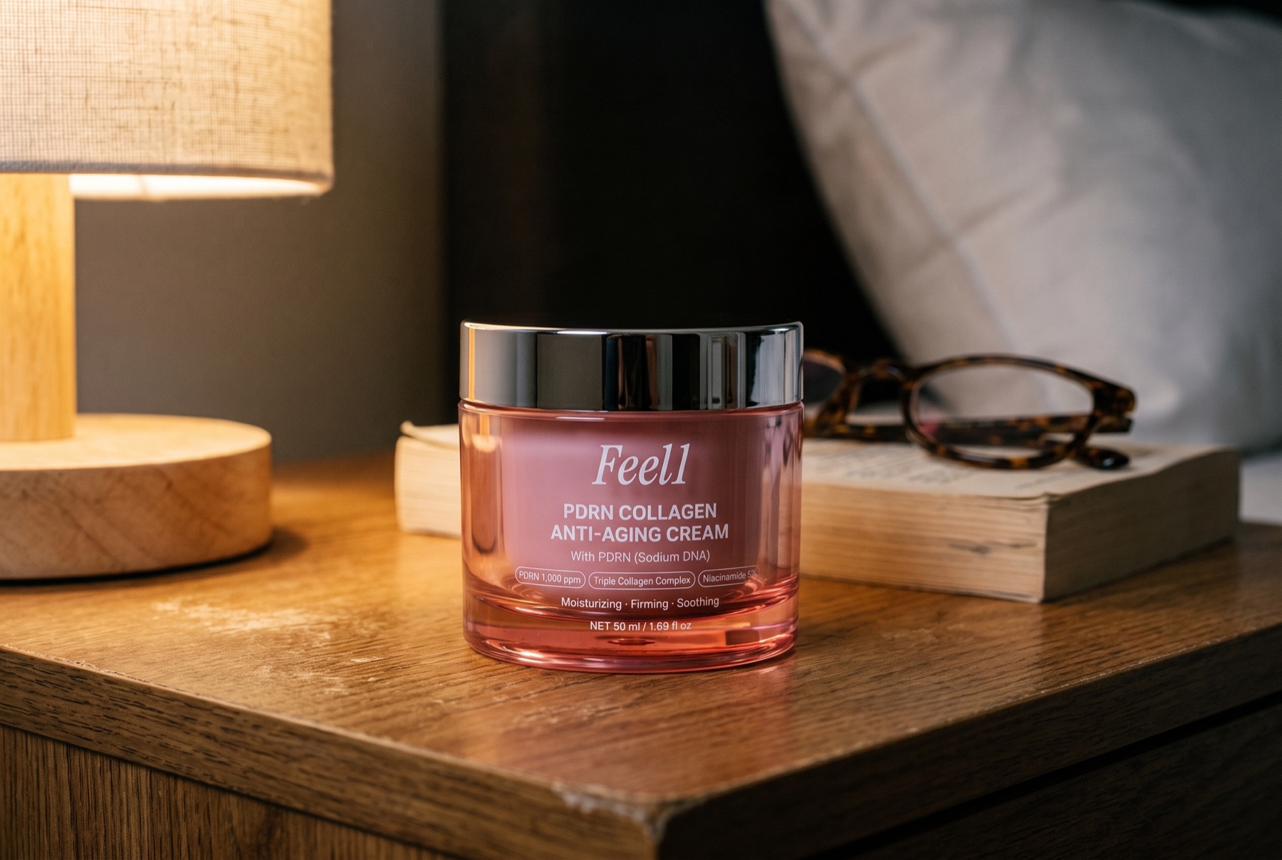 Feel1 PDRN Collagen Anti-Aging Cream, a pink glass jar with a chrome lid, on a wooden nightstand beside a paperback and reading glasses