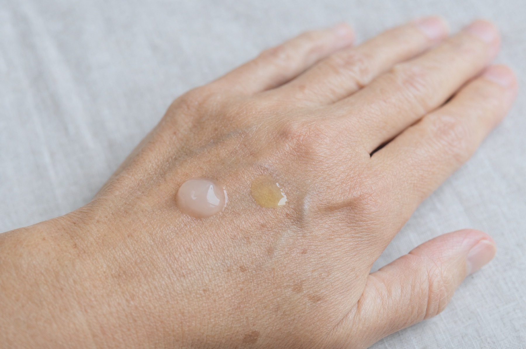 Back of a hand with a dab of pale pink gel cream next to a dab of yellowish retinol serum