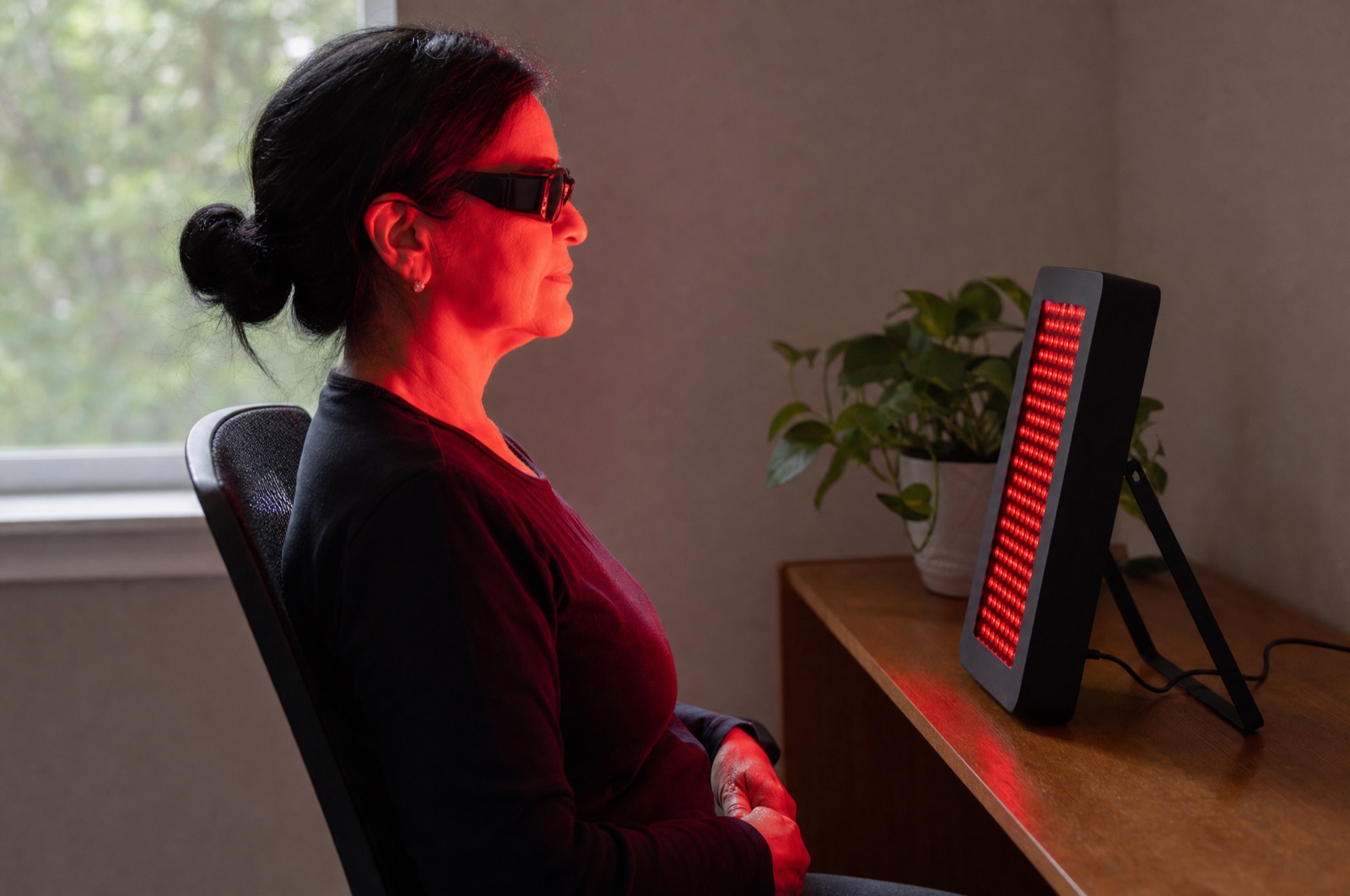 Woman in her late fifties wearing protective goggles in front of a tabletop red light panel at a home desk